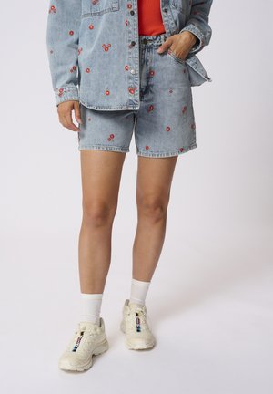 Set cu jachetă din denim și pantaloni scurți de culoare albastră deschisă, cu broderie florală roșie. Adidași albi cu un finisaj texturat și accente cu logo.