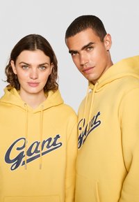 Gule hetter med en marineblå "Gant"-logo på brystet, med snorer og en frontlomme. Stoffet ser mykt og deilig ut.