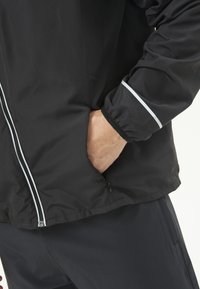 Personne portant une veste noire avec une bande réfléchissante sur la manche, la main dans la poche latérale zippée, et un pantalon noir assorti.