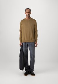 Un pull vert olive à manches longues avec un ourlet côtelé, associé à un jean en denim gris et des baskets noires. Portant un sac sombre.