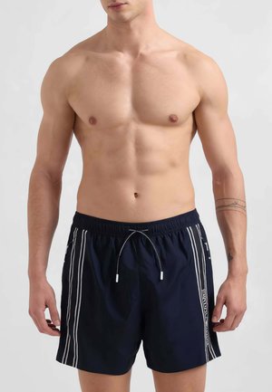 Man zonder shirt die marineblauwe zwemshorts met witte verticale strepen en zijzakken draagt, staand tegen een effen lichte achtergrond.