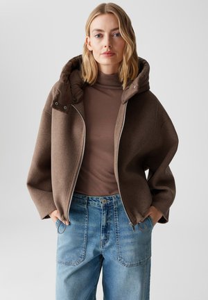 Donna bionda che indossa una giacca marrone con zip e colletto imbottito, dolcevita marrone e jeans a vita alta blu chiaro, con le mani in tasca, su uno sfondo neutro.