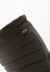 Bota de goma en verde oliva oscuro con superficie texturizada y un patrón en relieve. Presenta un puño de punto flexible y un pequeño acento en tres colores.