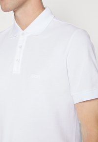 Weißes Polohemd mit klassischem Kragen, kurzen Ärmeln und einem dezenten Logo. Hergestellt aus glattem, leichtem Material mit einer schlichten Textur.