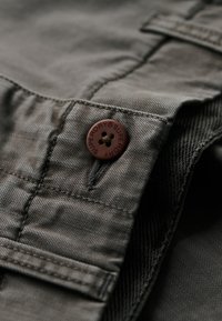 Tela gris con una textura suave, que presenta un botón marrón oscuro visible marcado "SUPERDRY" y costuras bien cosidas.