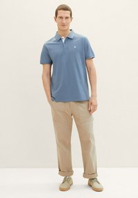 Hellblaues Poloshirt mit Kragen und dezentem Logo. Kombiniert mit beigen Cargohosen und hellen Schuhen, vor neutralem Hintergrund stehend.