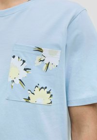 Lyseblå bomulds T-shirt med en blomster-mønstret brystlomme prydet med gule og hvide blomster samt grønne detaljer. Korte ærmer.