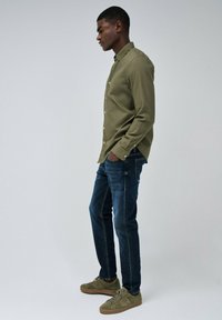 Camisa de botones verde oliva, vaqueros oscuros y zapatillas verde oliva. Corte slim, textura suave y detalles mínimos. Vista de perfil lateral.