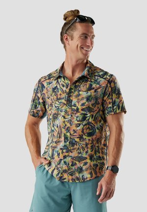 Mann mit Sonnenbrille auf dem Kopf, der ein buntes, kurzärmeliges Hemd mit Blumenmuster und türkise Shorts trägt, lächelt und seitlich wegschaut vor einem grauen Hintergrund.