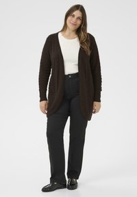 Brauner, gestrickter Cardigan mit einem V-förmigen Ausschnitt und gerippten Ärmeln. Tragen über einem weißen Oberteil, kombiniert mit schwarzen Hosen und Schuhen.