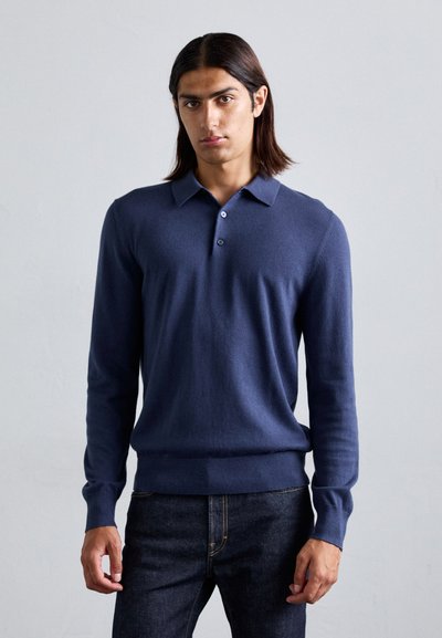 Polo suéter de manga larga en color marino, de tejido suave, con cuello, tapeta de tres botones y dobladillo acanalado. Combinado con jeans de mezclilla oscuros.