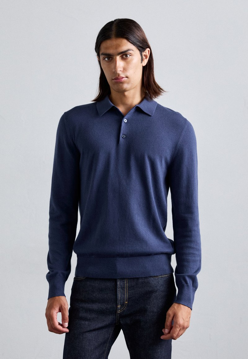 Navy long-sleeve polo sweater in soft fabric, featuring a collar, three-button placket, and ribbed hem. Worn with dark denim jeans.