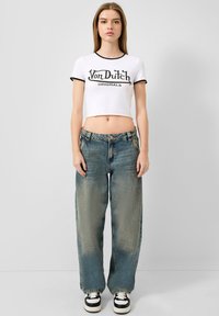 Biely skrátený tričko s čiernym lemom a textom "Von Dutch Originals", v kombinácii s voľnými bledomodrými džínsami a čiernymi teniskami.