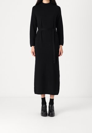 Woman wearing a long black knit dress with long sleeves and a belt, paired with black socks and black platform shoes, standing against white background.