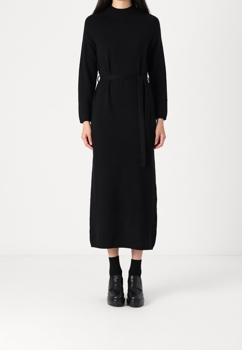 Femme portant une longue robe noire en tricot à manches longues avec une ceinture, associée à des chaussettes noires et des chaussures compensées noires, debout sur fond blanc.