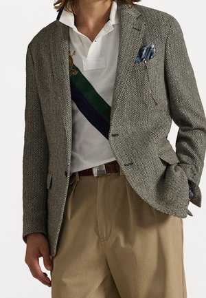 Homme portant un blazer beige à chevrons avec un mouchoir de poche bleu, un polo blanc avec une rayure diagonale verte et marine, et un pantalon kaki.