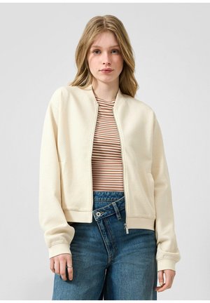Junge Frau mit langen blonden Haaren trägt eine cremefarbene, strukturierte Reißverschlussjacke über einem braun-weiß gestreiften Hemd und hoch taillierten blauen Jeans.