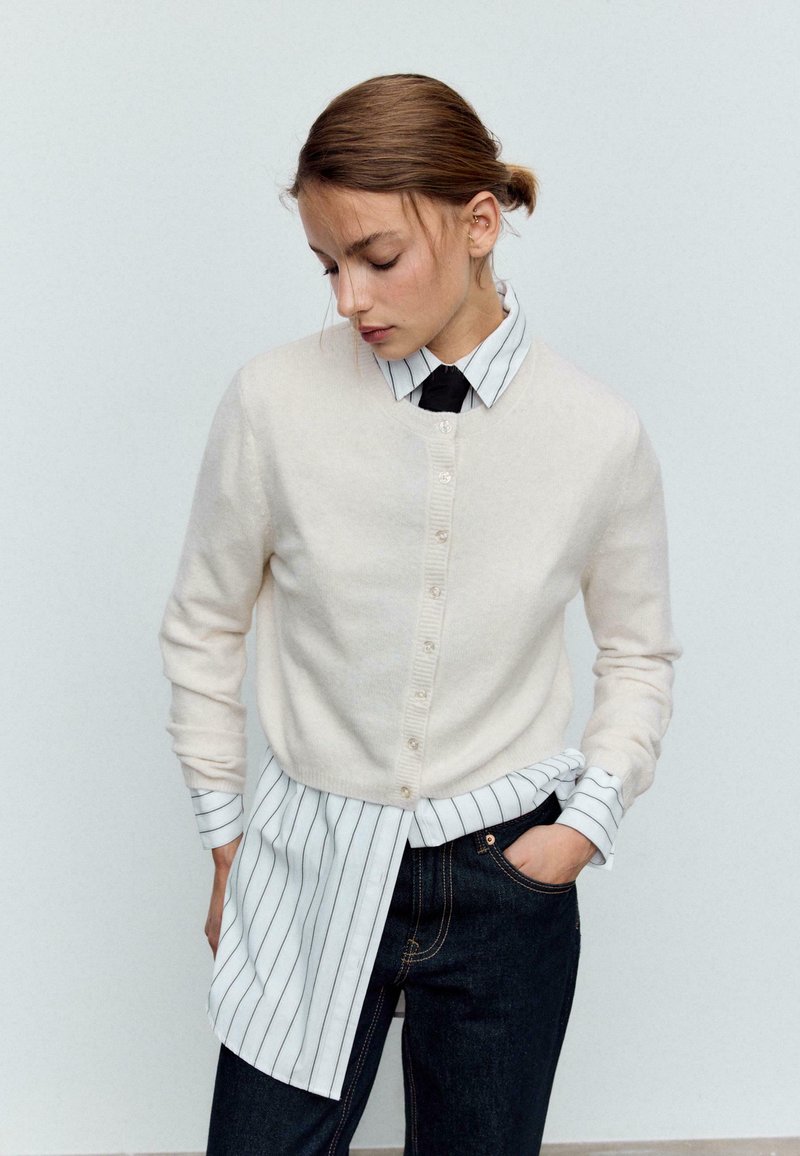 Cardigan beige clair en maille avec fermeture à boutons, porté sur une chemise rayée blanche et noire, assorti à un pantalon en denim foncé.