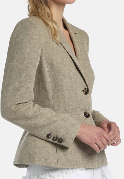 Mujer vestida con un blazer de lino beige con dos botones frontales y tres botones en los puños, combinado con una falda blanca con volantes.