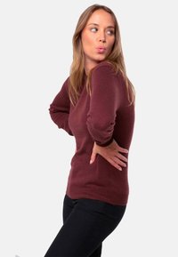 Pull bordeaux avec une texture douce, manches longues et col rond, associé à un pantalon noir. Vue de côté montrant la coupe ajustée.