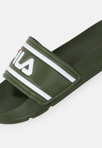 Olivgrön halksandaler i gummi med en texturerad yta; har ett vitt rand och en framträdande logotyp på remmen.