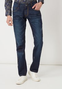 Vaqueros de denim azul oscuro con tachuelas decorativas en los bolsillos y los lados, con un acabado texturizado y sutiles detalles desgastados.