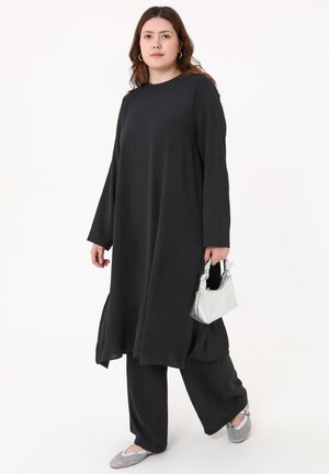 Donna con lunghi capelli castani che indossa un vestito nero largo e pantaloni, tiene una piccola borsa argentata e indossa scarpe basse argentate.