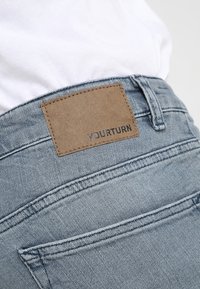 Jeans de mezclilla azul deslavado con una etiqueta de cuero marrón texturizado que dice "YOURTURN" en la parte posterior de la cintura, con bolsillos traseros estándar y costuras.