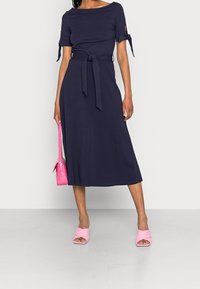 Robe bleu marine à manches courtes, avec des liens latéraux et une ceinture à la taille ; associée à des sandales plates roses et un sac à main rose texturé.