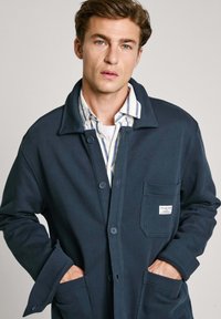 Marinblå bomulljacka med krage, knappstängning, bröstficka och synliga sömdetaljer. Under jackan bärs en randig skjorta.