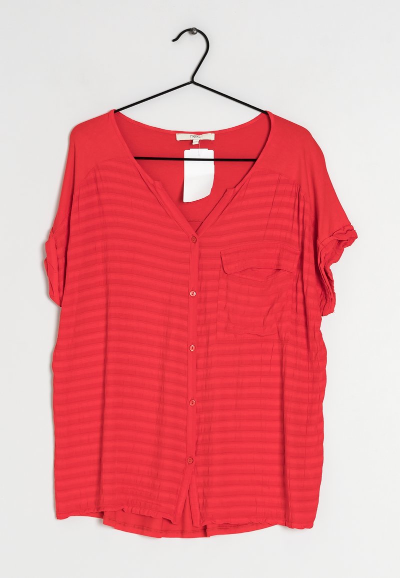Blusa corta rossa a maniche corte con scollo a V, chiusura con bottoni, motivo a coste testurizzate e una singola tasca sul petto. Appesa a un gancio nero.
