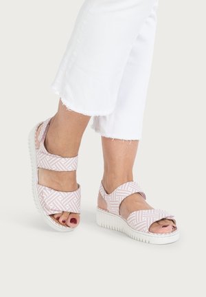 Füße in weißen, gekürzten Hosen, die rosa-weiße Sandalen mit dicken weißen Sohlen tragen, vor einem einfarbigen Hintergrund.