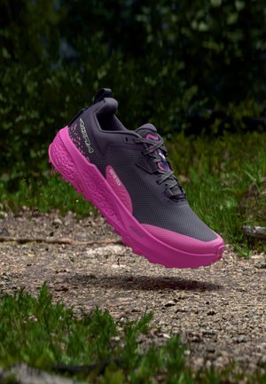 Mørkegrå og lys pink vandresko med GORE-TEX og Vibram-sål svæver over en grusstier i en grøn udendørs omgivelser.