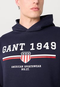 Tengerészkék kapucnis pulóver, nagy fehér felirattal: "GANT 1949" és egy pajzs logóval, piros és fehér csíkokkal. Puha, textúrált anyagú, állítható kapucnival.