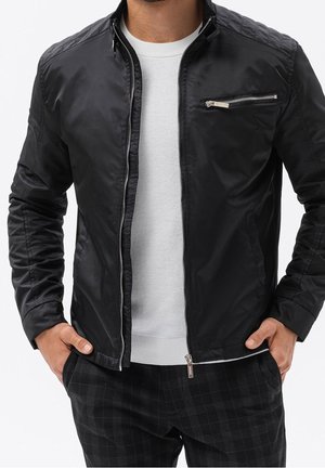 Homme portant une veste noire zippée sur une chemise blanche texturée et un pantalon noir à carreaux, les mains dans les poches.