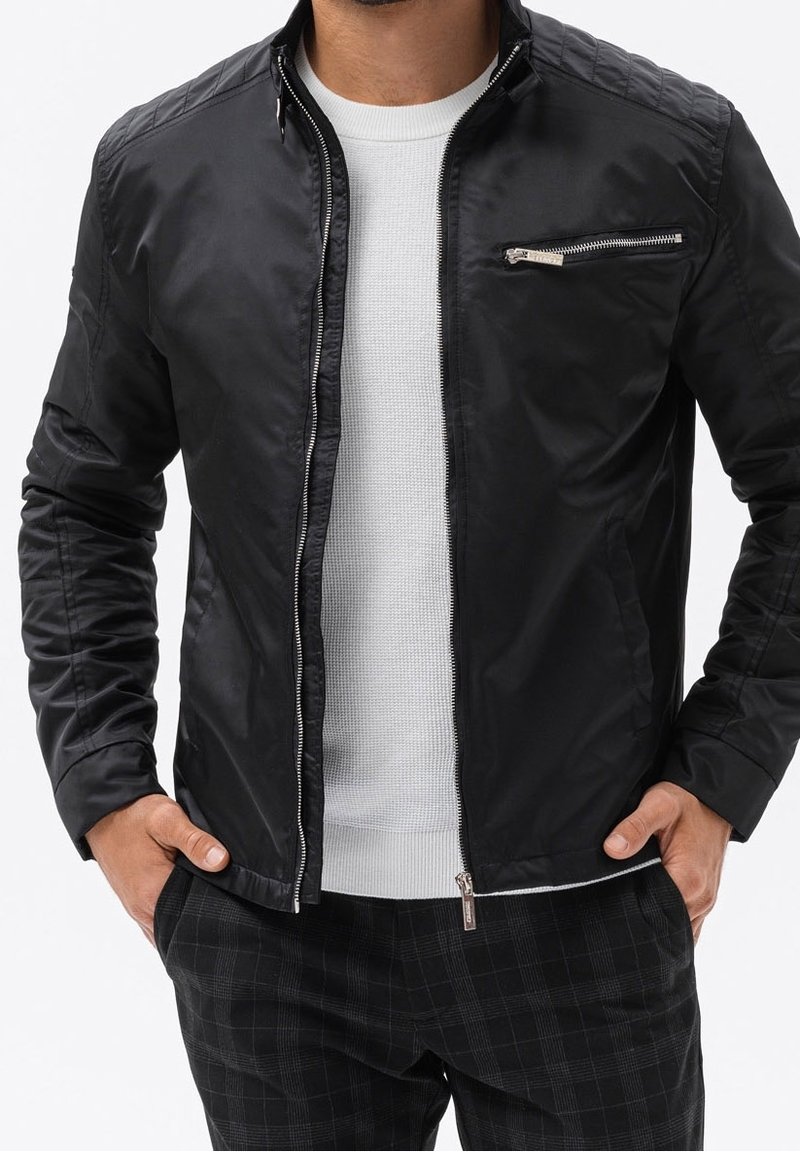 Homme portant une veste noire zippée sur une chemise blanche texturée et un pantalon noir à carreaux, les mains dans les poches.