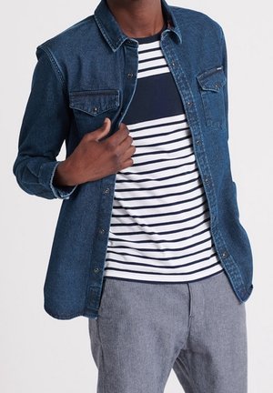 Mand iført en blå denimskjorte over en hvid og marineblå stribet t-shirt med grå bukser, hånd justerer skjortekraven.