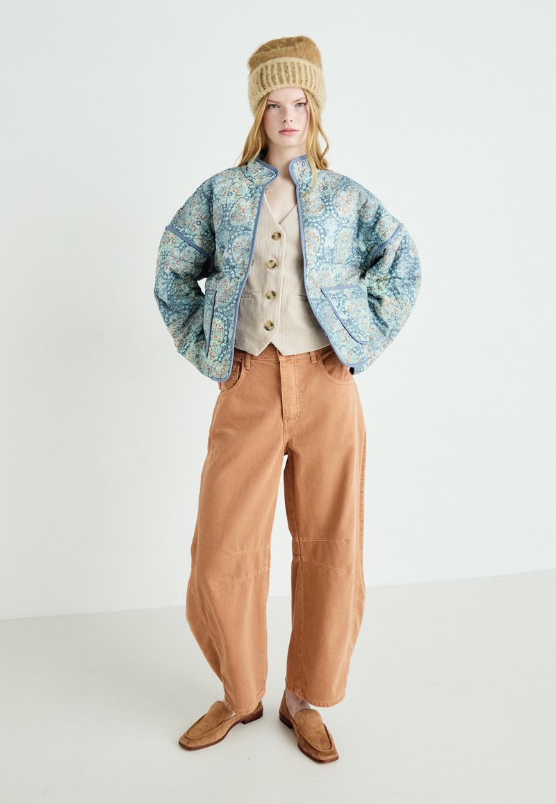Gewatteerde jas met blauwe bloemenprint, beige vest met knopen, rustkleurige wijde broek en tan instap schoenen. Zachte texturen overal.