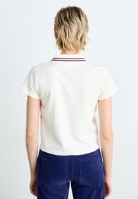 Polo à manches courtes, couleur crème, avec une texture côtelée et un col rayé bordeaux. Porté avec un pantalon en velours côtelé bleu marine, offrant une coupe décontractée.