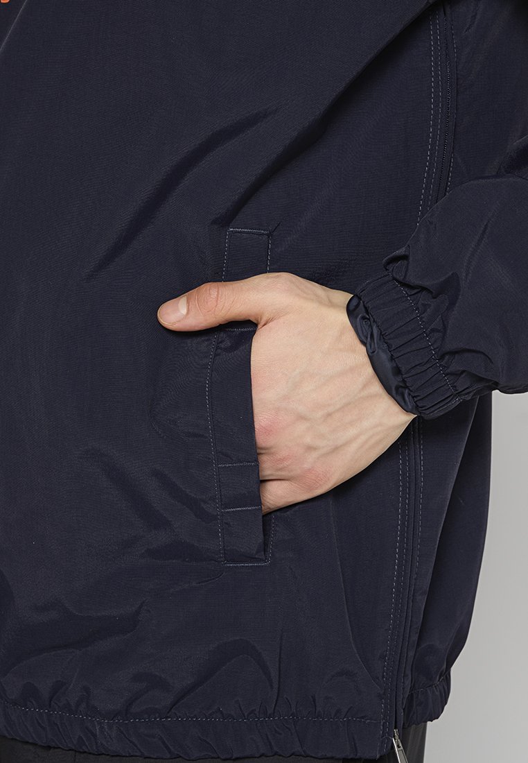 Chaqueta ligera de color azul marino con puños elásticos, bolsillo lateral y una textura suave con un detalle de costura mínimo. Dobladillo ajustado.