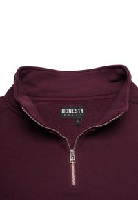 Burgundin värinen vetoketjullinen huppari, jossa on korkea kaulus, hopeinen vetoketju ja sisäpuolella fleecekangas. Etiketissä lukee "HONESTY" ja koko "L."