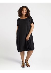 Schwarzes Kurzarmkleid mit lockerem Schnitt und rundem Ausschnitt. Glattes, leichtes Material mit gerafften Details am Saum. Schwarze Sandalen mit goldenen Akzenten.