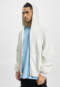 White zip-up hoodie in soft fabric with a large hood and front pockets, layered over a light blue t-shirt. Minimalist design, elastic cuffs.