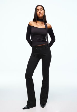 Frau steht mit den Händen hinter dem Rücken, trägt ein schwarzes schulterfreies Oberteil mit Choker-Detail, schwarze enge Hose und schwarze spitze Schuhe.