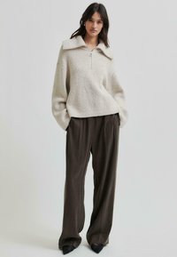 Gruby sweter w kremowym kolorze z zamkiem przy kołnierzu, zestawiony z brązowymi spodniami o szerokich nogawkach i czarnymi spiczastymi butami. Sweter ma wyraźny wzór splotu.