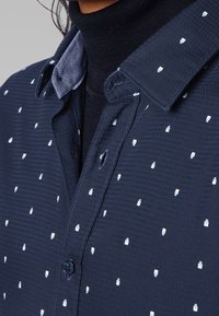 Marineblauwe shirt met een gestructureerde stof, versierd met een klein, herhalend geometrisch patroon in lichtblauw en wit. Details van de button-down kraag zijn zichtbaar.