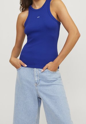 Woman wearing a fitted blue sleeveless top with JJXX logo and light blue high-waisted denim jeans with hands in pockets.