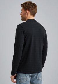 Camisa negra de manga larga con una sutil textura a rayas, que presenta un collar y un pequeño detalle metálico en la parte trasera. Llevada con jeans azul claro.