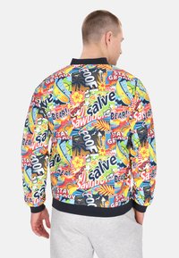 Bunte Bomberjacke mit verschiedenen grafischen Drucken, darunter Text und tropische Motive. Hergestellt aus leichtem Stoff mit einem schwarzen Kragen.
