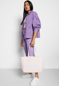 Veste en denim violet et jean assorti, accompagnés d'un sac fourre-tout rose clair avec un logo bleu. Baskets blanches avec des détails texturés.
